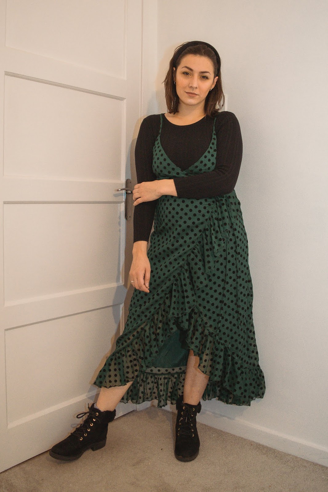 I am standing with my left arm holding my right. I am wearing a black ribbed long sleeve top under a green frill polka dot dress that is a mid length. I am wearing black boots with fluffy trim.