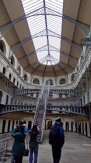 Oval shaped interior of a building. Roof is comprised of windows looking out onto a grey sky. Four levels of creme colored walls with black jail cell doors. Stairs in the center of the room go directly to the fourth level. Blue grey dark steel cages surround the stairs and each level of the jail cells