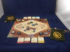 A game of War Chest in progress. The board has several tokens around it, with the draw bags nearby, several piles of tokens around it, and four cards on each side.