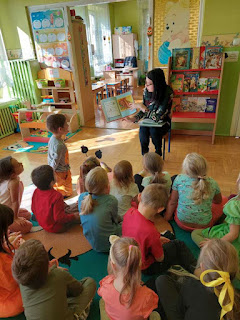 tło: sala przedszkolna. Pani bibliotekarka pokazuje dzieciom siedzącym na dywanie książkę o jesieni.