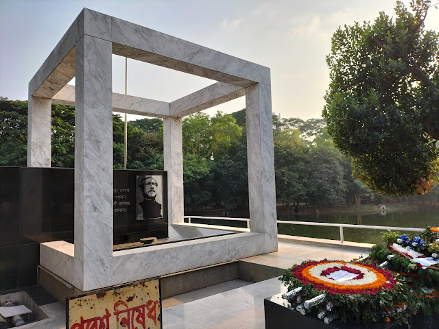 Monument of a large cube frame with picture of Sheik Mujibur Rahman and dedication in the middle, and flower arrangements placed in front of it
