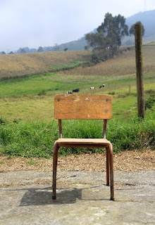 Old school wooden chair with metal frame positioned on bare ground in front of a wooden post. Grass-covered rolling hills, a large tree and cows in the background.