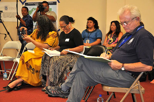 several adults seated in folding chairs in a conference room, all taking notes, with a man with a camera in the background