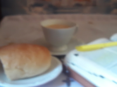 Cantinho Café com Deus em uma mesa com uma xícara de café, uma bíblia aberta com uma marca de texto em cima e um prato com um pão caseiro