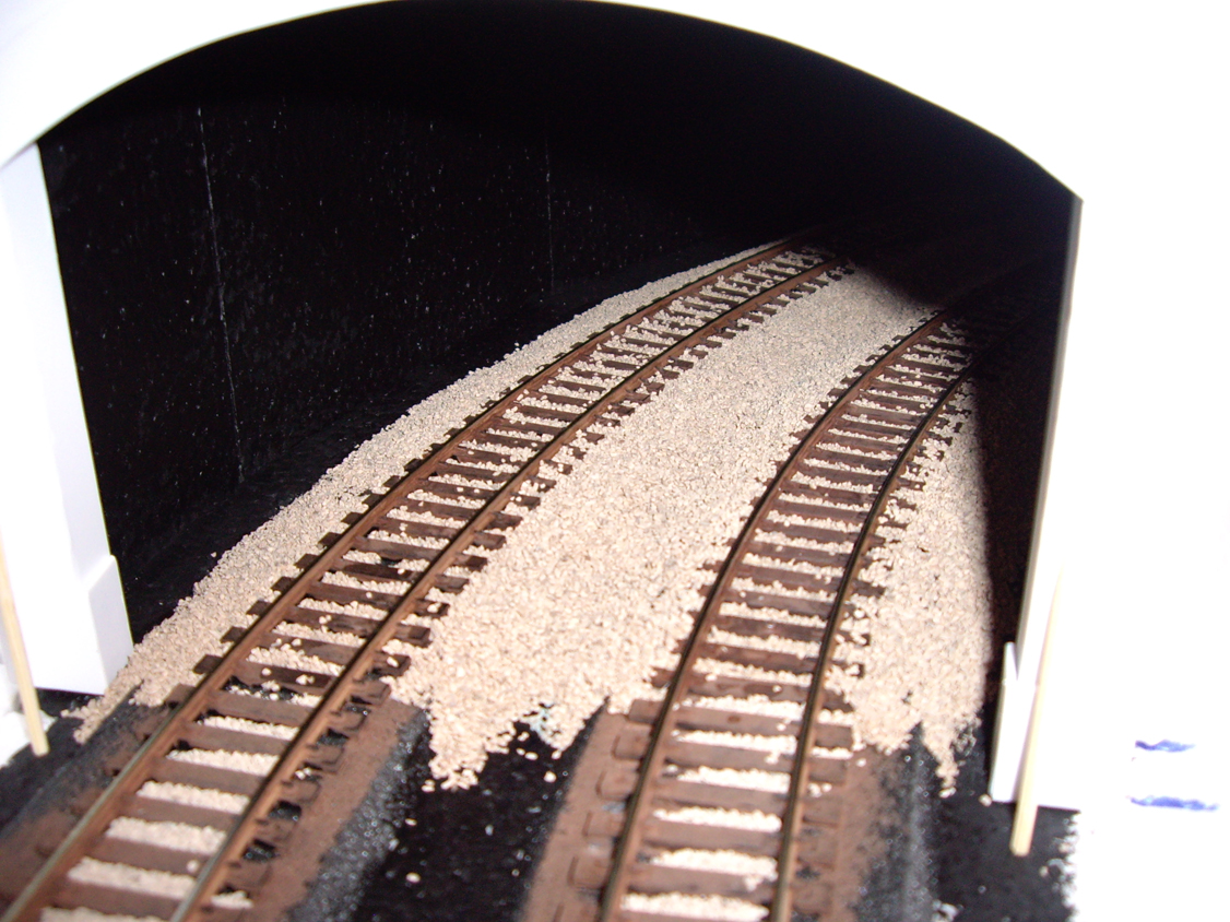 A set of half ballasted model railroad tracks coming out of a styrene tunnel portal