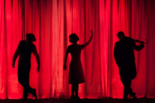 a silhouette of three performers behind a red theatre curtain 