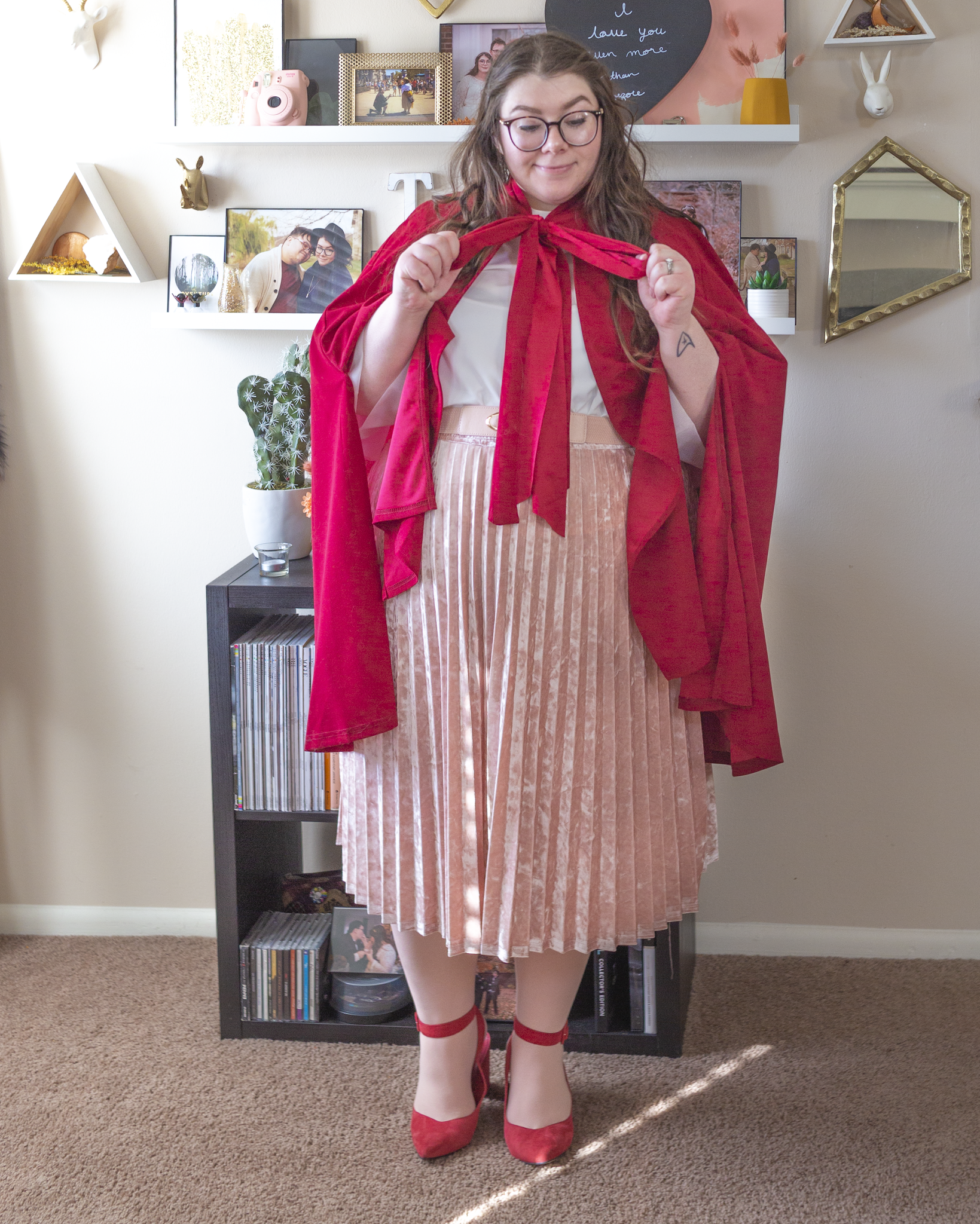 An outfit consisting of a red cape tied into a bow around the neck over a white high neck blouse, tucked into a pastel pink velvet pleated midi skirt and red ankle strap heels.