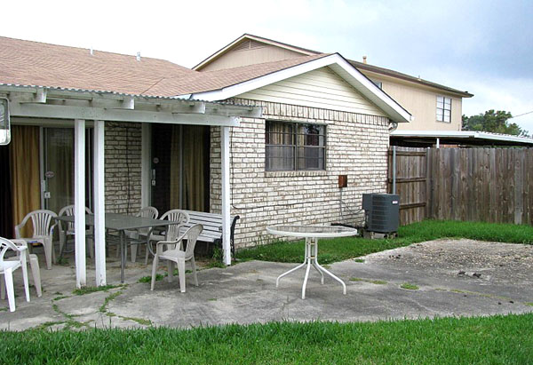 the back yard of a humble-looking house of white-painted brick, with a small poured concrete patio and a few pieces of patio furniture