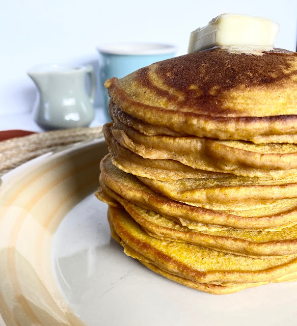 A stack of pumpkin pancakes on a plate with syrup and coffee mug in the background.