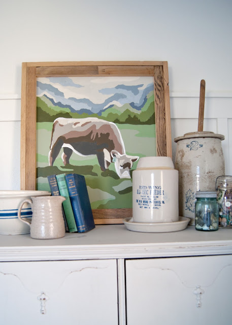 Farmhouse Master Bedroom Reveal - Benjamin Moore Simply White, Paint by number cow painting . antique crocks, red wing, chicken feeder, books, mason jars