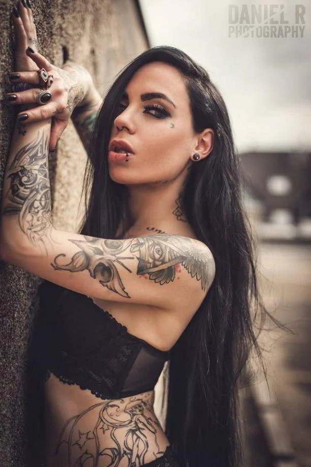 Chica morena apoyada en un muro, lleva tatuaje del ojo que todo lo ve en su brazo