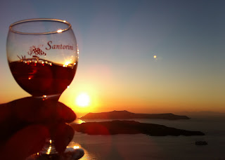 Holding a glass of Vinsanto with the sun setting over the Santorini volcano in the caldera.