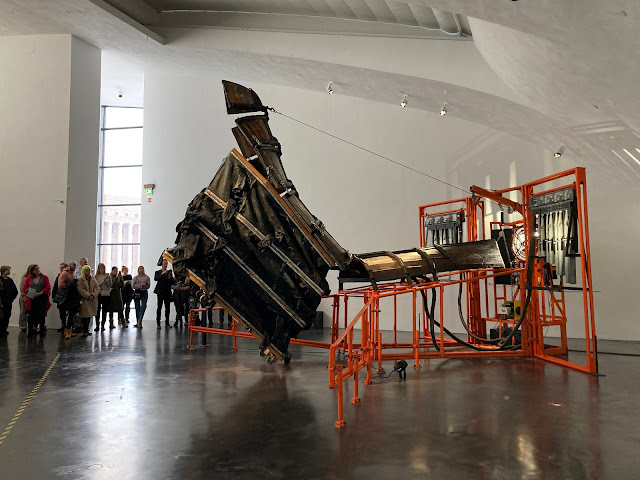 A large sculpture with an orange metal frame and an upside down wooden boat rears up in a gallery, as a line of people watch.