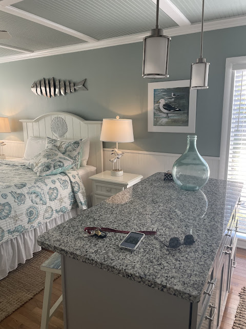 Another photo of the place we stayed at. This picture shows the queen size bed with an ocean themed light blue and white bed spread with a nightstand. On top of the night stand is a beach themed lamp. The picture also shows the black and white granite counterop of the island in the kitchen. the ceiling is painted a gray blue with white beams running in a perpendicular and parallel pattern. There is a silver fish accent piece hanging above the bed. There is also a window to the right of the nightstand.