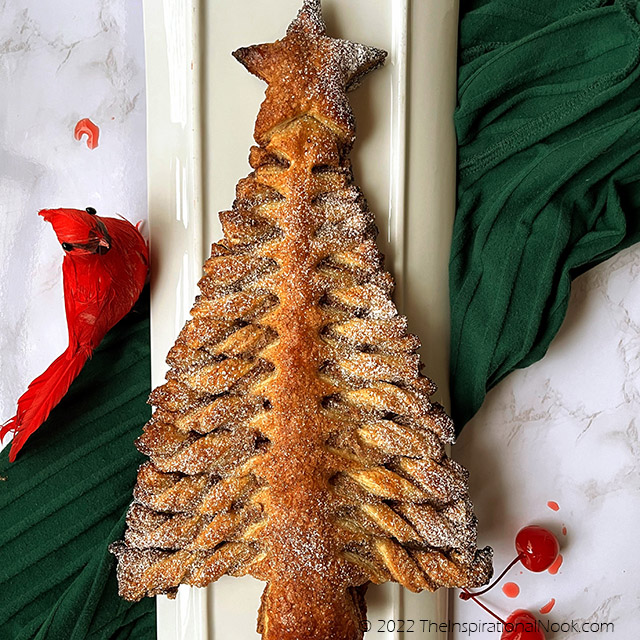 Pull apart Christmas tree puff pastry dessert decorated with powdered sugar and glace cherries on a white plate, with a red cardinal bird sitting on a dark green cloth