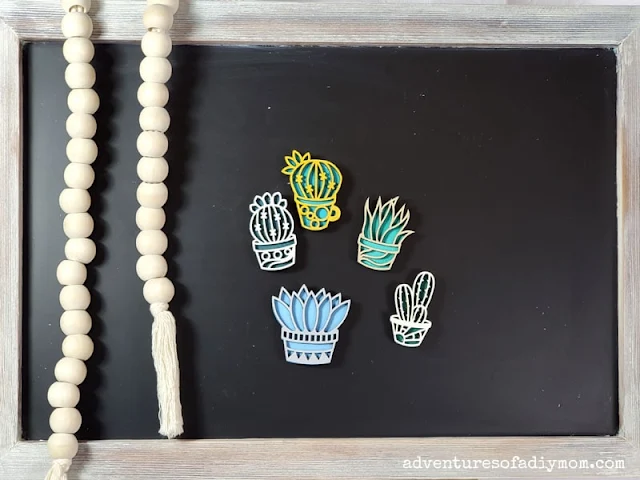 5 handmade succulent magnets cut with the xTool S1 laser cutting machine hanging on a chalkboard with a string of wood beads dangling next to them.