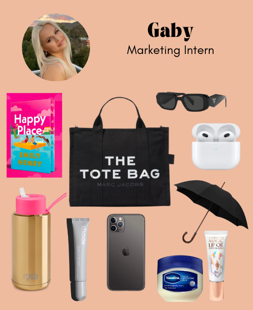Image of marc jacobs black bag with book, sunglassses, airpods, lip balm, vaseline, rhode lip gloss, waterbottle, phone and umbrella surrounding it. They are all against a pale orange backdrop.