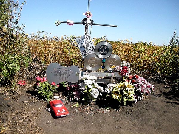 roadside shrine; a cross made of steel pipe, engraved records and metal cut-out guitar, numerous flowers, a small car model