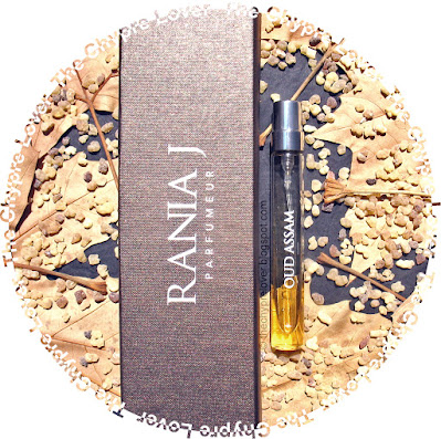 The bottle and brown box of Oud Assam by Rania J. (eau de parfum) lying flat on a dark gray table amongst brown dried sycamore leafs and incense granules