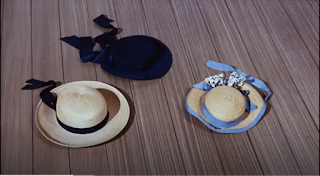 Three hats on a wooden floor. It is from a scene in Mary Poppins. There is a black hat and two straw hats. One straw hat has blue ribbon and flowers the other has a black ribbon.