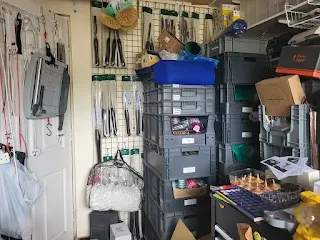 Compact workshop with labeled grey Euro containers stacked for storage, wall-mounted grid displaying woodturning tools, and various supplies including ribbons, packaging materials, and finished wooden products