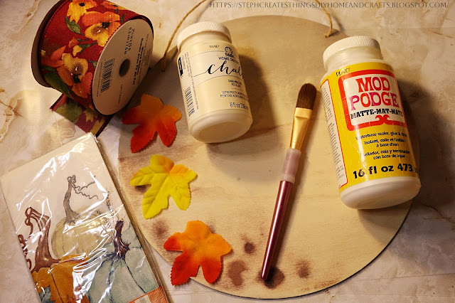 Craft materials are displayed on a table. Ribbon, decorative napkins, faux fall leaves, Mod Podge, and acrylic paint.