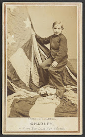 An old daguerreotype photo depicting a light-skinned boy with European features. A large American flag is draped off to the left of the image, covering the floor and the stool the boy is sitting on. Under the photo the following has been typed: Freedom's Banner, CHARLEY, A Slave Boy from New Orleans.