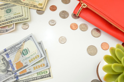 A pack of currencies on a table. Dollars notes, gold and silver coins, a pink wallet and a potted plant.