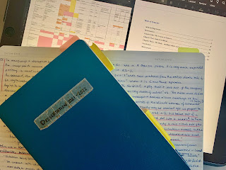 a blue notebook sitting on top of handwritten notes with a colour coded spreed sheet and word document in the background.