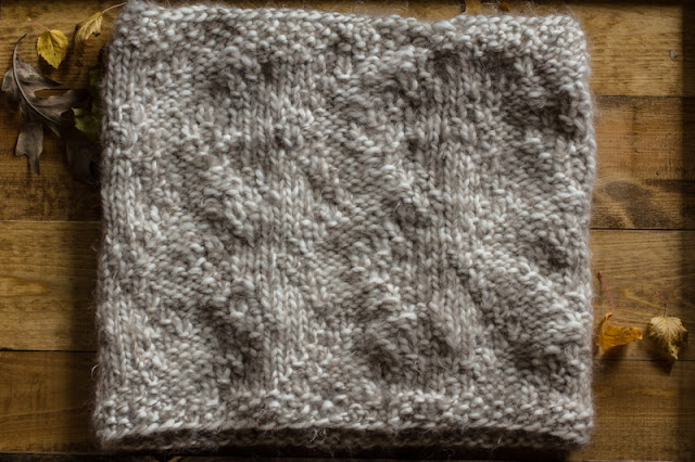 Flat show of A cream and tan knit soft cowl on a wood surface next to dried leaves. The arrow pattern is slightly noticeable.