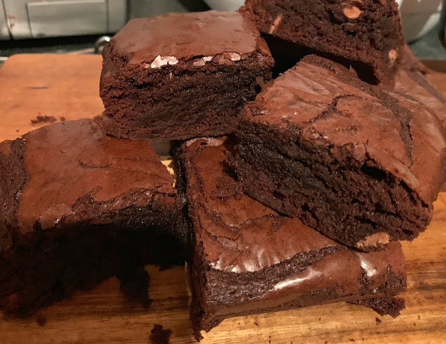 picture shows 6 chocolate brownies, and a brown wooden chopping board. The brownies are moist and have a shiny, papery top, and chunks of giant chocolate buttons through them