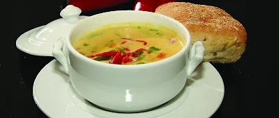 Eine Schüssel Suppe mit einem Brötchen. Das Bild zeigt eine weiße Schüssel mit einer cremefarbenen Suppe, in der rote und grüne Gemüsestücke und Kräuter zu sehen sind. Die Schüssel steht auf einem weißen Teller, auf dem dem auch ein goldbraunes, frisch gebackenes Brötchen liegt. Der Hintergrund ist schwarz, was die weiße Schüssel und den Teller hervorhebt. Das Bild wirkt realistisch und appetitlich.
