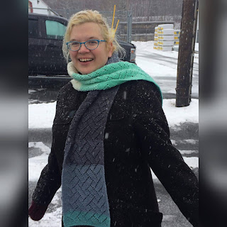 A lace scarf in a gradient yarn of several shades of mint and gray is careless draped on a blonde woman in a black coat laughs as snow falls.