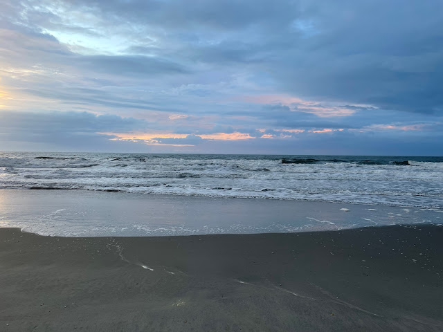 A gray sky over the ocean. The waves seem to be coming in roughly. The picture is taken from the wet, sandy beach.