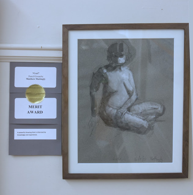 Pencil and gouache drawing of nude woman, seated, with face mask, hanging on gallery wall. Labels on left side say "Cori" Pencil/gouache Matthew Mattingly Merit Award A powerful drawing that is infused with knowledge and experience .