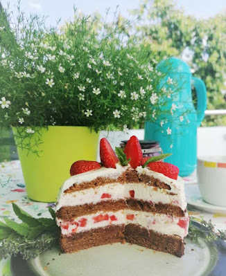 Erdbeer-Mascarpone-Torte-Anschnitt zeigt zwei drittel der Torte auf dem Teller im Anschnitt, so dass die einzelnen Schichten aus Boden, Cremes und Erdbeerstücken zu sehen sind. Das ganze auf einem für den Kaffee gedeckten Tisch auf einem Balkon bei schönem Wetter. Oben auf der Torte sieht man noch die ganzen Erdbeeren und 2 Blätter einer Zitronnenmmelisse.