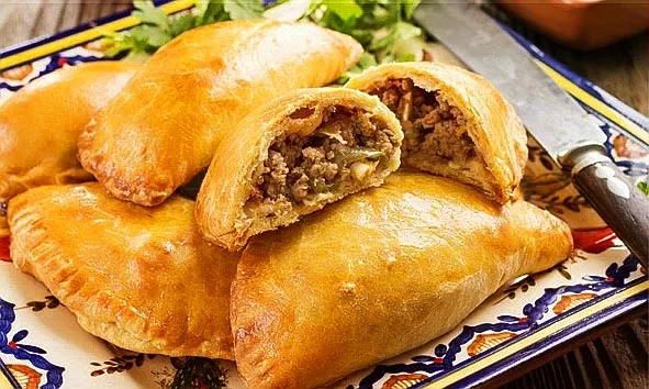 A platter of golden-baked meat fatayer, showcasing triangular pastries filled with a flavorful mixture of ground beef or lamb, onions, pine nuts, and a blend of Middle Eastern spices such as allspice, cinnamon, and cumin. These savory hand pies feature a crisp, flaky crust enveloping a juicy, aromatic filling, making them a beloved appetizer or snack across Lebanese, Syrian, Palestinian, and broader Levantine cuisines. Often served warm with sides like yogurt or tahini sauce, meat fatayer are a staple of traditional Middle Eastern home cooking and festive gatherings. This image captures the inviting texture and rich flavors of authentic meat fatayer.