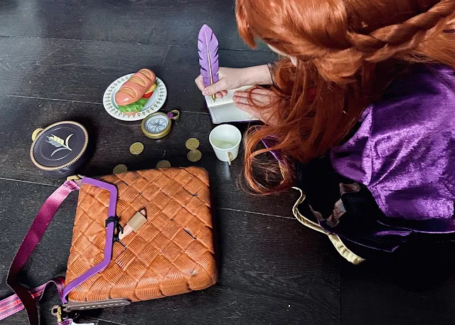 Overhead photo of a girl dressed as Anna from Frozen 2 with the shopDisney range. She is writing in a notepad and has the contents of the plastic travel bag in front of her including money, a water bottle, mug, plates, sandwich, compass, coins and quill pen