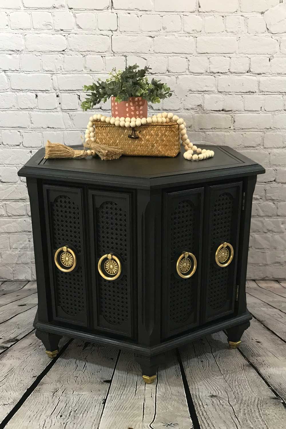 Finished black chalk painted vintage mid-century hexagonal end table with polished original hardware staged with wicker box, plant and wooden beads