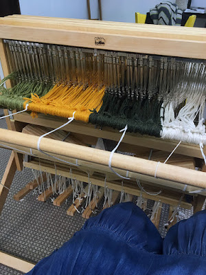 Three-quarter view of a loom from the back right corner. Light green, marigold, dark green, and white wool yarn are knotted onto the rear apron rod in sequence; the apron rod is a few inches from the metal heddles, supported by white cords at either end. The viewer's knees in a denim skirt are visible in the lower right of the photo.