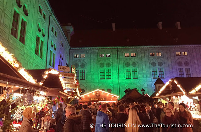 People walk on a square between illuminated wooden stalls decorated with Christmas ornaments in front of a large period-style property illuminated in green.