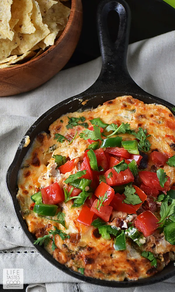 Delicious Jalapeno Popper Dip in a sizzling cast iron skillet, garnished with a colorful medley of diced tomatoes, jalapeno, and fresh cilantro. Perfectly paired with a bowl of crispy chips for a mouthwatering snack.