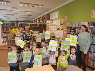 Dzieci prezentują swoje prace plastyczne wraz z przedszkolanką i paniami bibliotekarkami. Na tle regały z książkami oraz tablica korkowa z wiosennymi zdjęciami.