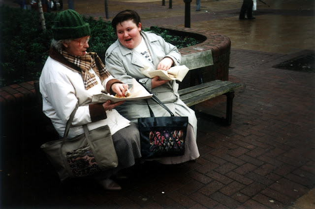 Two women sat chatting on a bench with their fish and chips. The older lady is wrapped up in a coat, scarf and woolly hat, and the younger lady is wearing her coat open