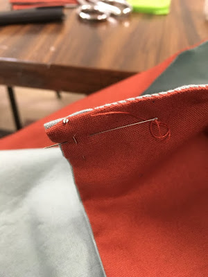 A close-up of the edge of a piece of red fabric lined with grey-blue, with a pin holding the turned-in edges together at the corner, and a red-threaded needle stuck in the fabric just past the existing whip stitches over the edge.