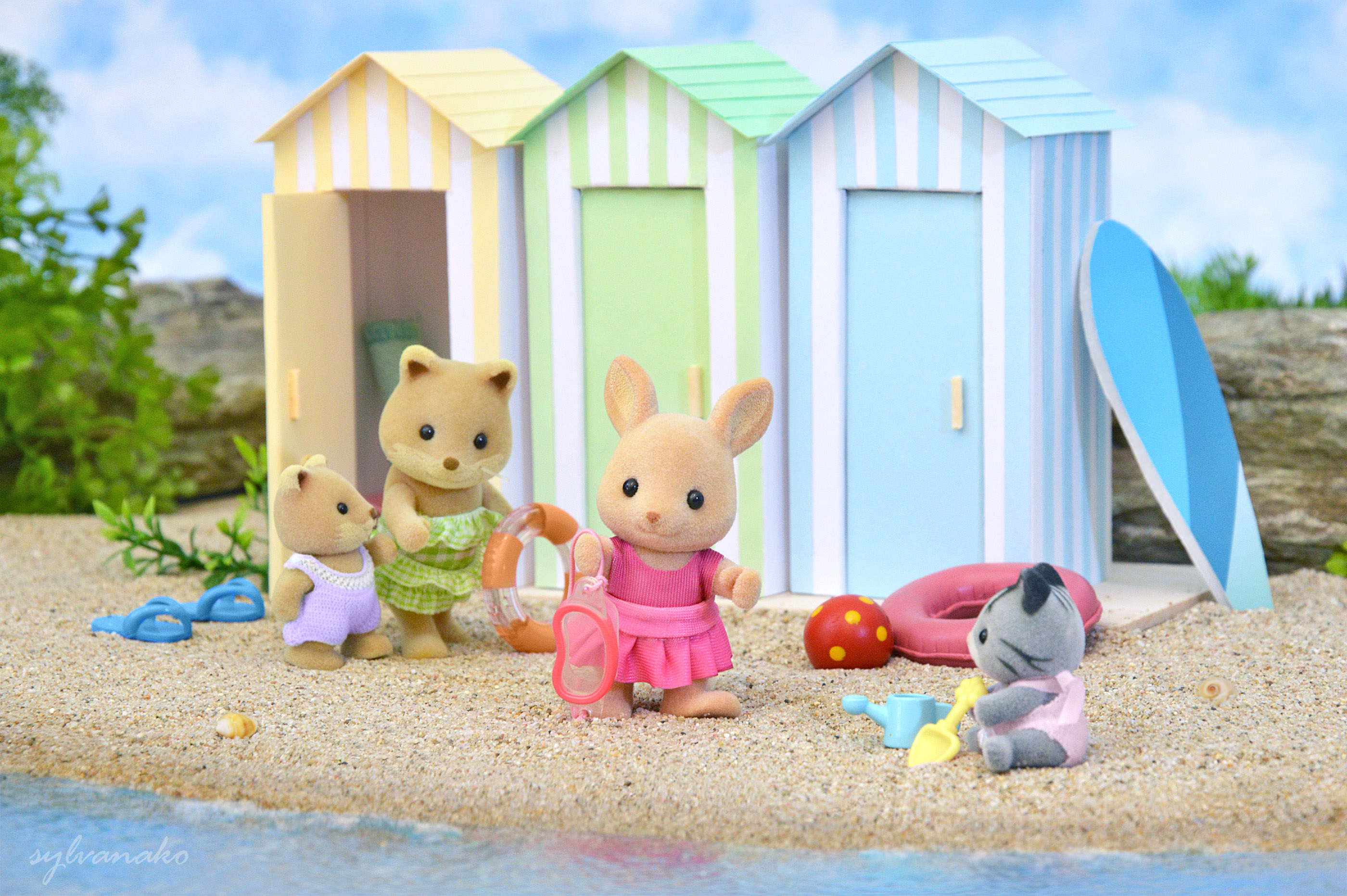 Calico Critters on a sandy beach in bathing suits, in front of beach pastel changing rooms.