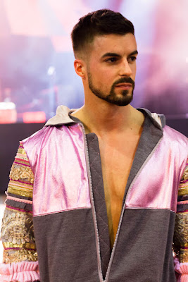 Homme mannequin qui défile en portant une veste à bandes rose et grise. Les manches sont aussi des bandes de tissus assemblés, roses, grises et or.