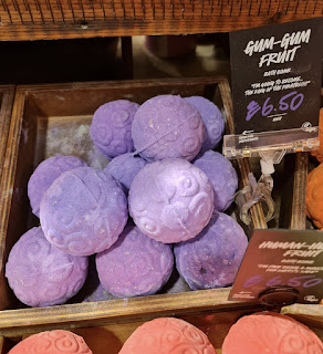 A photo of a light wooden light brown square box filled with purple spherical bath bombs Wirth swirled indents in it next to a black card that says gum-gum fruit bath bomb in white font on a bright background