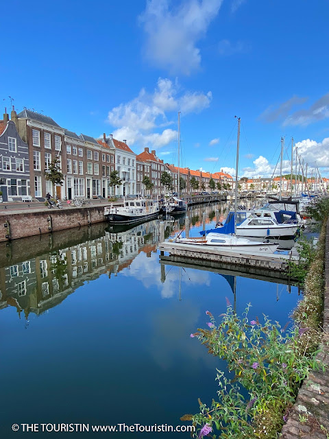 Reflections of the sky and white and blue mid-sized yachts in a harbour lined by a long row of three-storeyed red brick period houses.