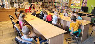 sala biblioteczna. Pani bibliotekarka siedzi na przeciwko grupy przedszkolaków, siedzącej przy bezowych drewnianych stolikach i czyta im legendę o Lechu, Czechu i Rusie.